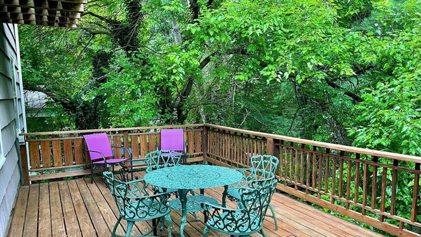 A wooden deck with a round table and chairs, surrounded by a wooden railing and lush greenery.