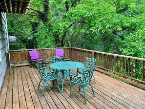 Una terraza de madera con una mesa redonda y sillas, rodeada por una barandilla de madera y abundante vegetación.