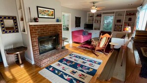 A living room with a brick fireplace, wooden flooring, a colorful rug, a pink sofa, a television, and a ceiling fan.