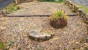 Una petita casa de pedra amb teulada inclinada, envoltada per un camí de grava i un jardí amb una banca de fusta i una planta en test.