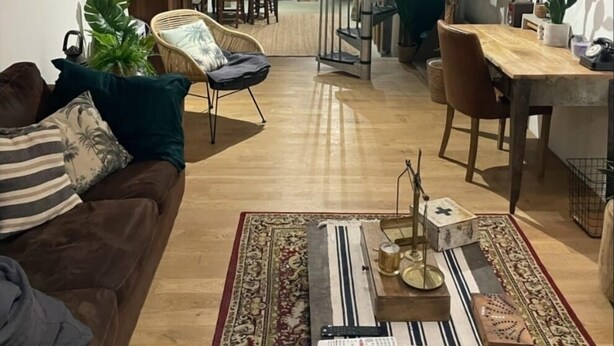 Un saló acollidor amb sòl de fusta, un sofà, una taula de centre amb un objecte decoratiu, i una escala que condueix a una zona de menjador amb taula i cadires de fusta.