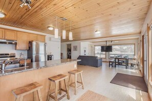 Een moderne keuken met houten kasten, een granieten aanrechtblad en roestvrijstalen apparatuur. Er staan houten krukken bij een groot keukenblok met barstoelen. Het plafond is van hout met hangende pendellampen.