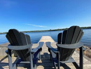 Twee Adirondack stoelen gericht op een houten steiger die uitloopt in een kalm meer, met een heldere lucht.