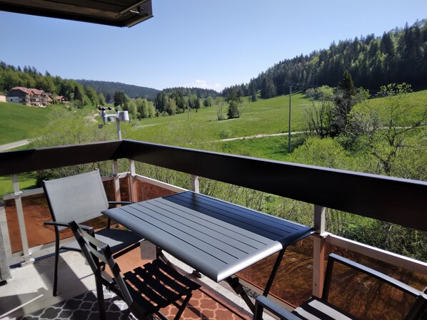 Ein Balkon mit Tisch und Stühlen, mit Blick auf eine grüne Landschaft mit Bäumen und einem Gebäude in der Ferne.