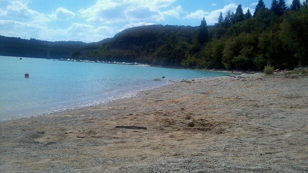 A beach with a clear view of the water, a sandy shore, and a forested hillside in the background. - Insertion Dans la Nature (Lamoura)
