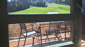 A balcony with a table and chairs, overlooking a green landscape with trees and a hill. - Insertion Dans la Nature (Lamoura)