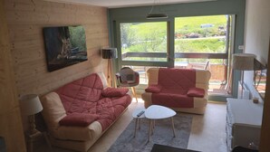 A cozy living room with a wooden wall, a red sofa, a white armchair, and a small table. There is a window with a view of greenery outside. - Insertion Dans la Nature (Lamoura)