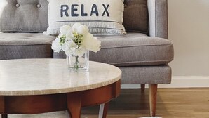 A grey sofa with a pillow that reads "RELAX" and a small vase with white flowers on a round wooden coffee table. - Urban Escape: Trendy 1-Bed Condo with Balcony (San Diego)