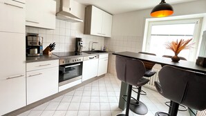A modern kitchen with white cabinets, a built-in oven, a coffee machine, and a dining area with a black table and bar stools. - Hirschtraum im Zentrum von Braunlage (Braunlage)