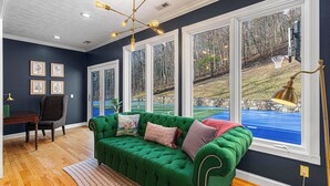 A room with a green sofa, a wooden floor, a desk with a chair, and large windows with a view of a blue pool and trees. - 7k SqFt Estate - Epic Amenities & Fun - Sleeps 20 (Biltmore Forest)
