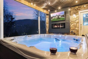 A hot tub with two glasses of red wine, a stone fireplace, and a TV on the wall.