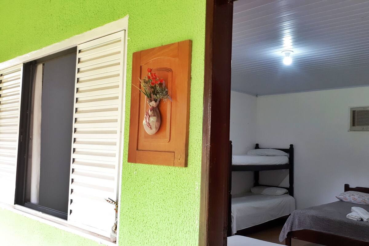 A room with a bunk bed, a single bed, a window with blinds, a wall-mounted air conditioning unit, and a ceiling light.