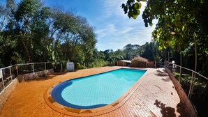 Piscina retangular com deck revestido de azulejos, cercada por uma grade de metal e árvores.