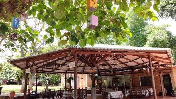 Uma área de refeições coberta ao ar livre, com mesas e cadeiras, rodeada por árvores e vegetação.