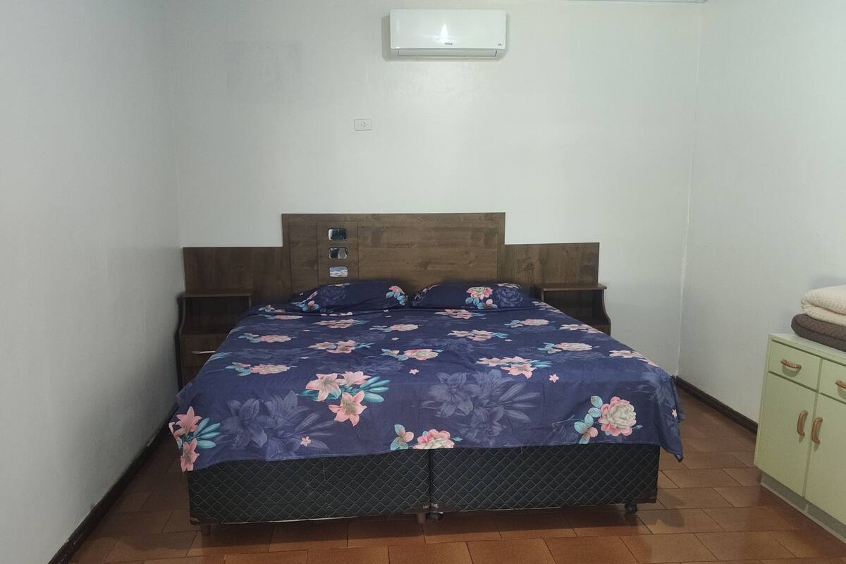 A bedroom with a bed, wooden headboard, nightstands, a small cabinet, and a wall-mounted air conditioner.