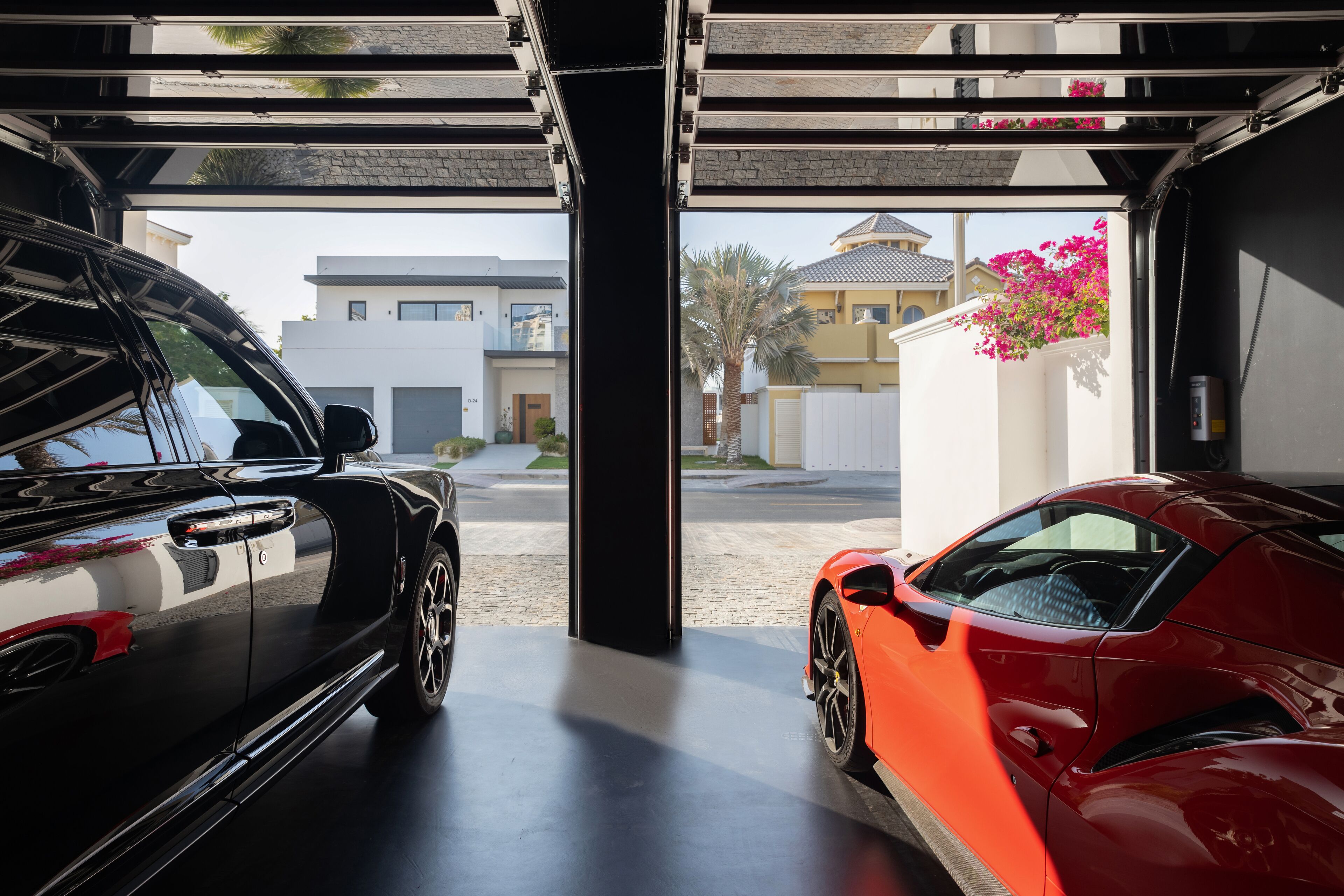 A garage with two cars, one black and one red, and a view of a residential area with houses and a palm tree.