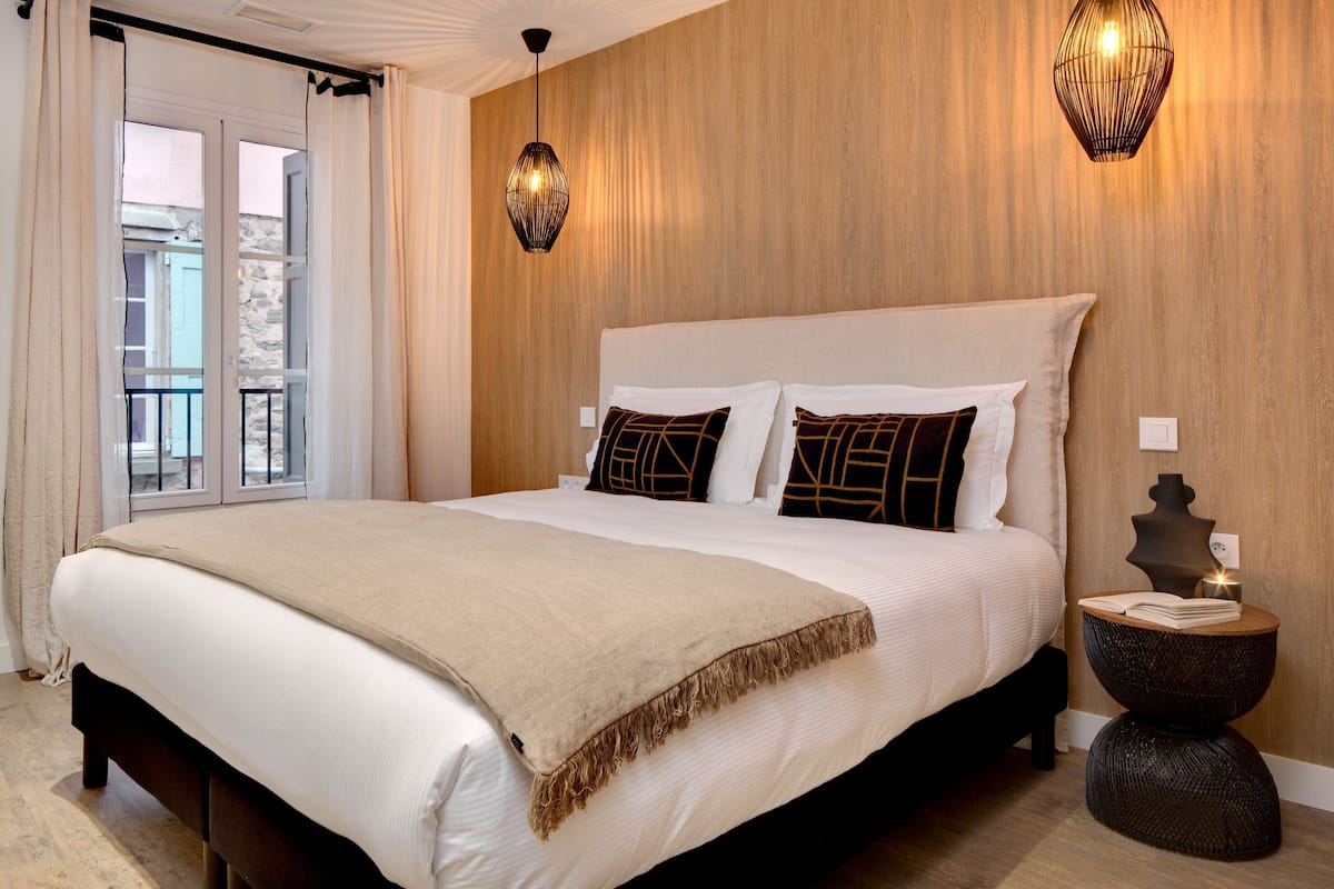 A hotel room with a large bed, two wall sconces, a bedside table with a book, and a view of the outside.