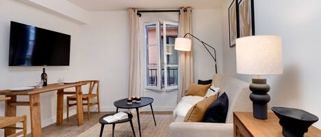 A modern living room with a flat-screen TV, wooden furniture, a sofa, a lamp, and a view of a balcony through a window.