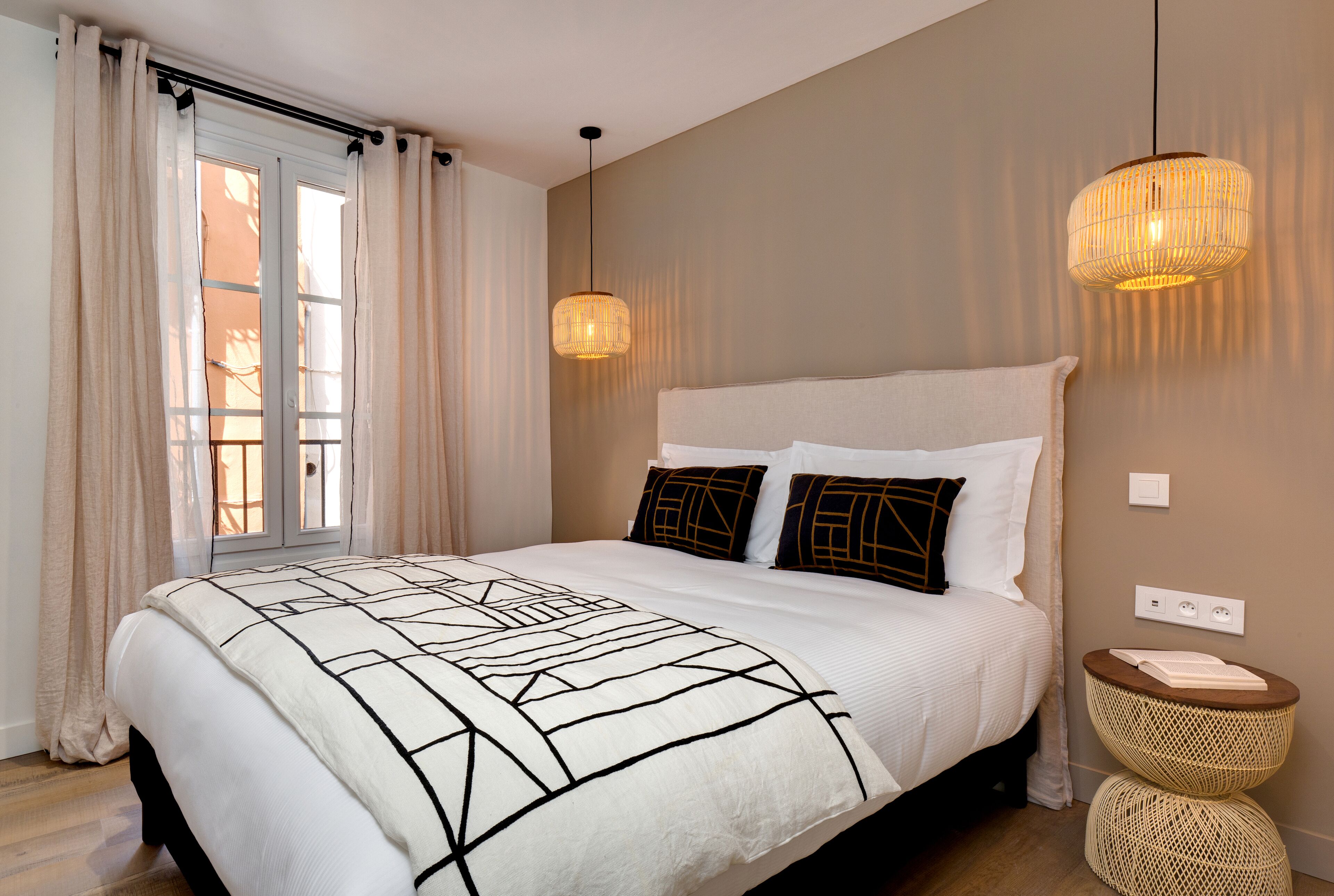Un lit bien fait, recouvert d’une couverture à motifs géométriques, une table de chevet avec un panier en osier, et une lampe suspendue.