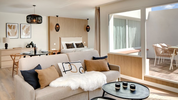 Una habitación de hotel moderna con sofá, mesa de comedor, cama y balcón con mobiliario de exterior.