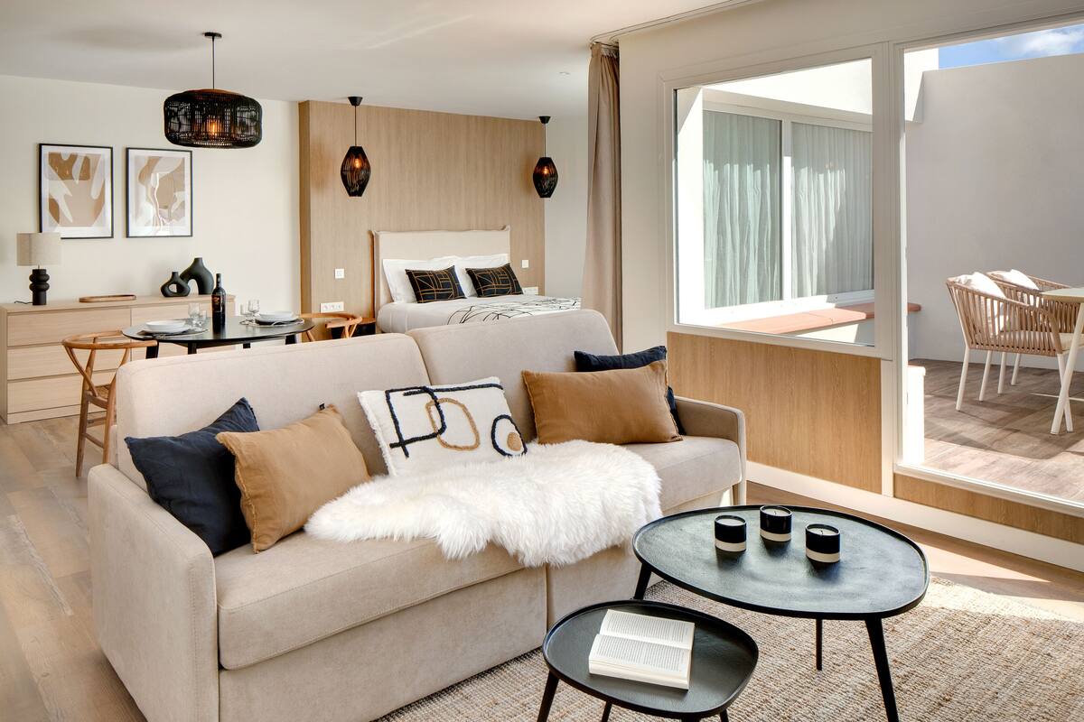 A modern hotel room with a sofa, a dining table, a bed, and a balcony with outdoor seating.