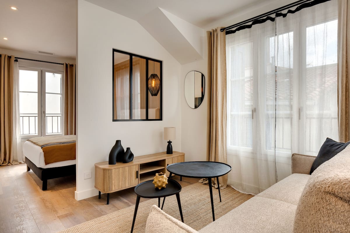 A modern hotel room with a bed, a sofa, a small table, a mirror, and a window with sheer curtains.