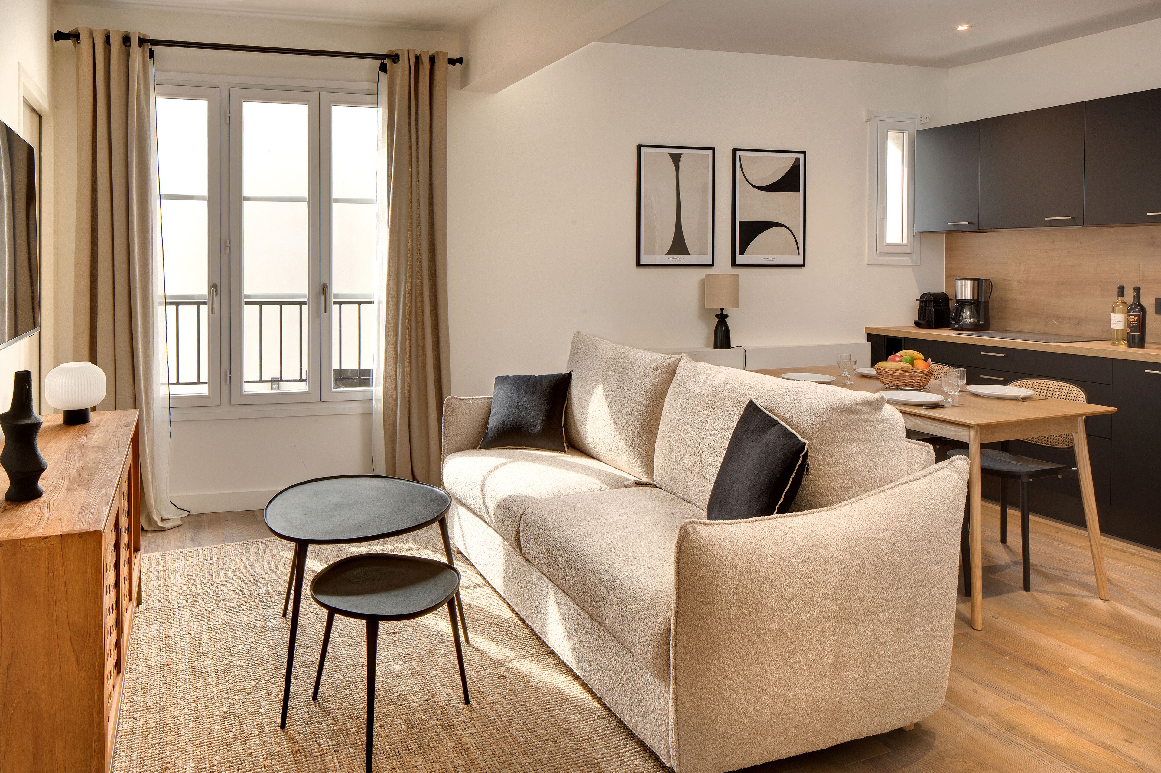 Un salon moderne avec un canapé beige, deux tables d’appoint noires, une table basse en bois et un coin cuisine doté de meubles sombres et d’un plan de travail en bois.