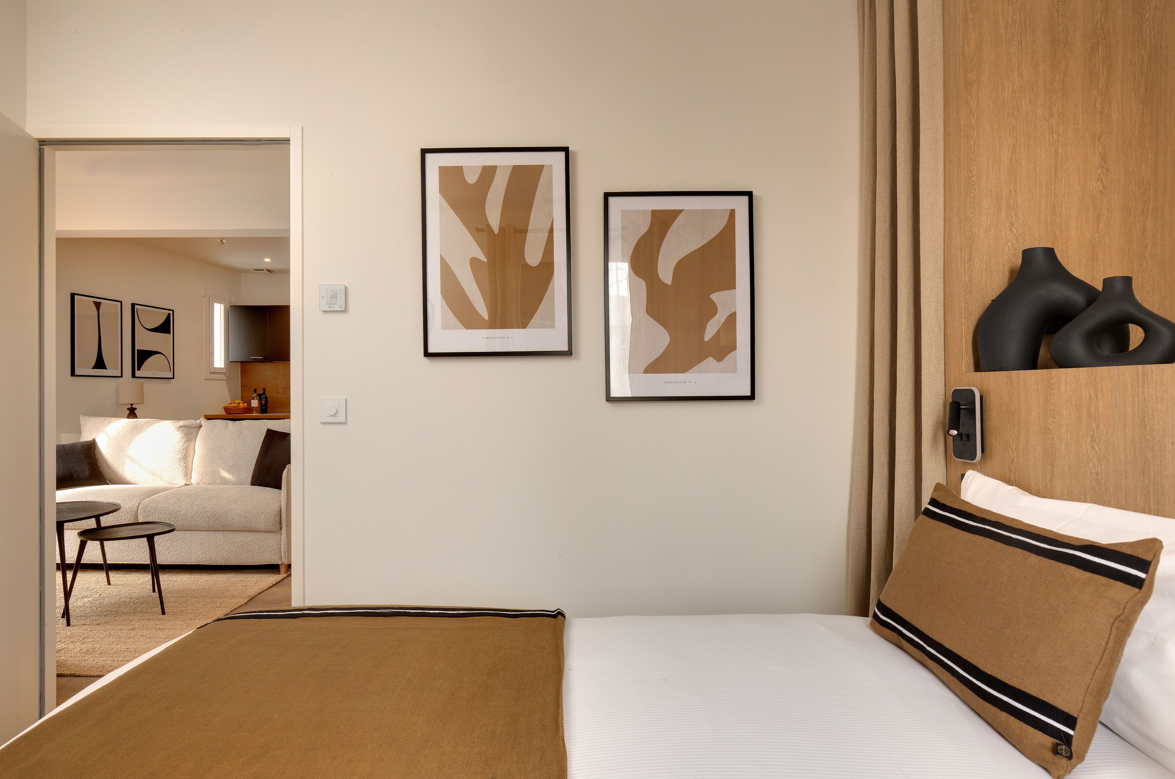 Habitación de hotel con una cama, un plaid marrón, dos cuadros abstractos enmarcados, un cabecero de madera y una pequeña mesa con una silla.