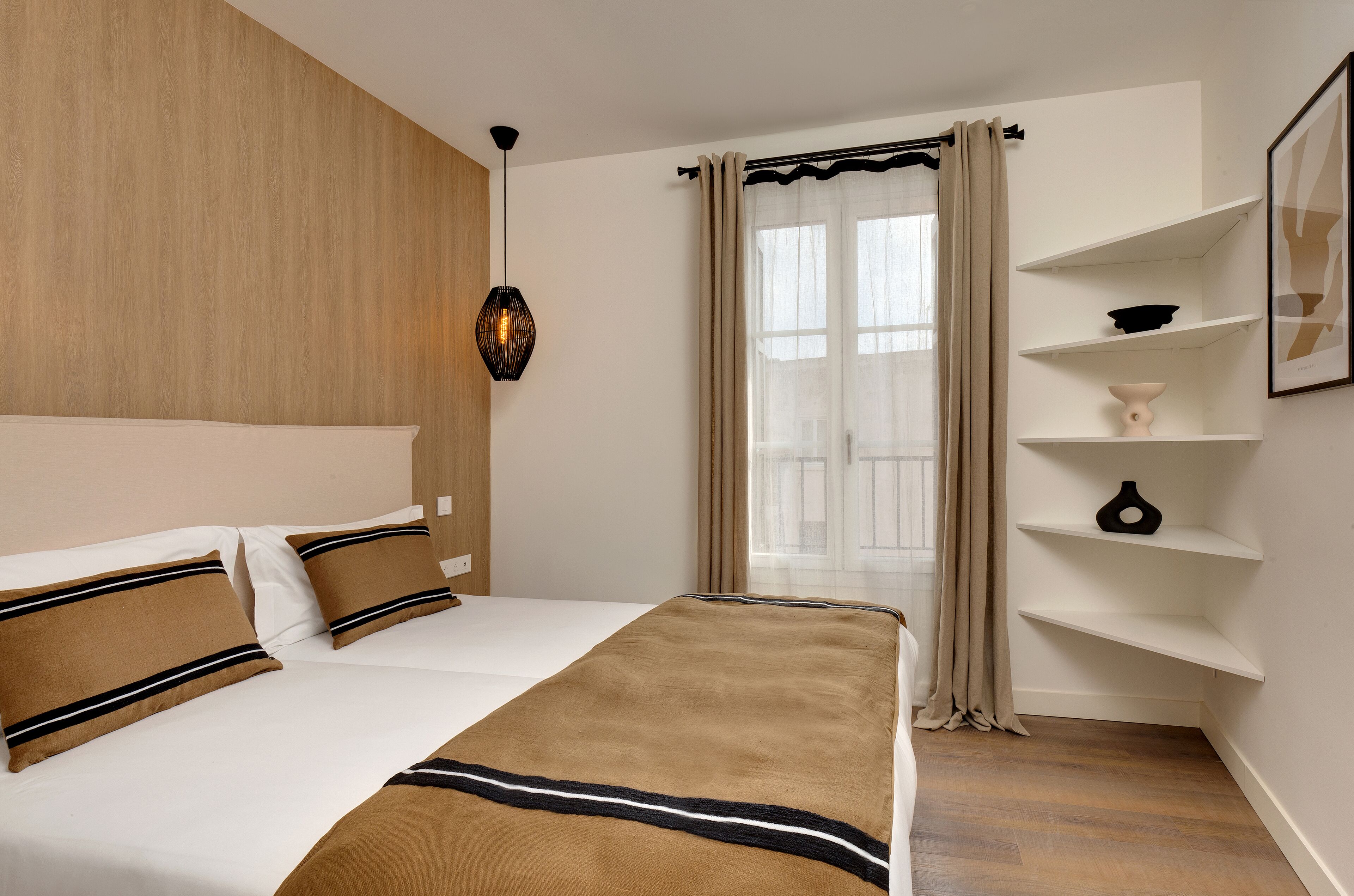 A bedroom with a large bed, a wooden headboard, a wall-mounted shelf with decorative items, and a window with curtains.