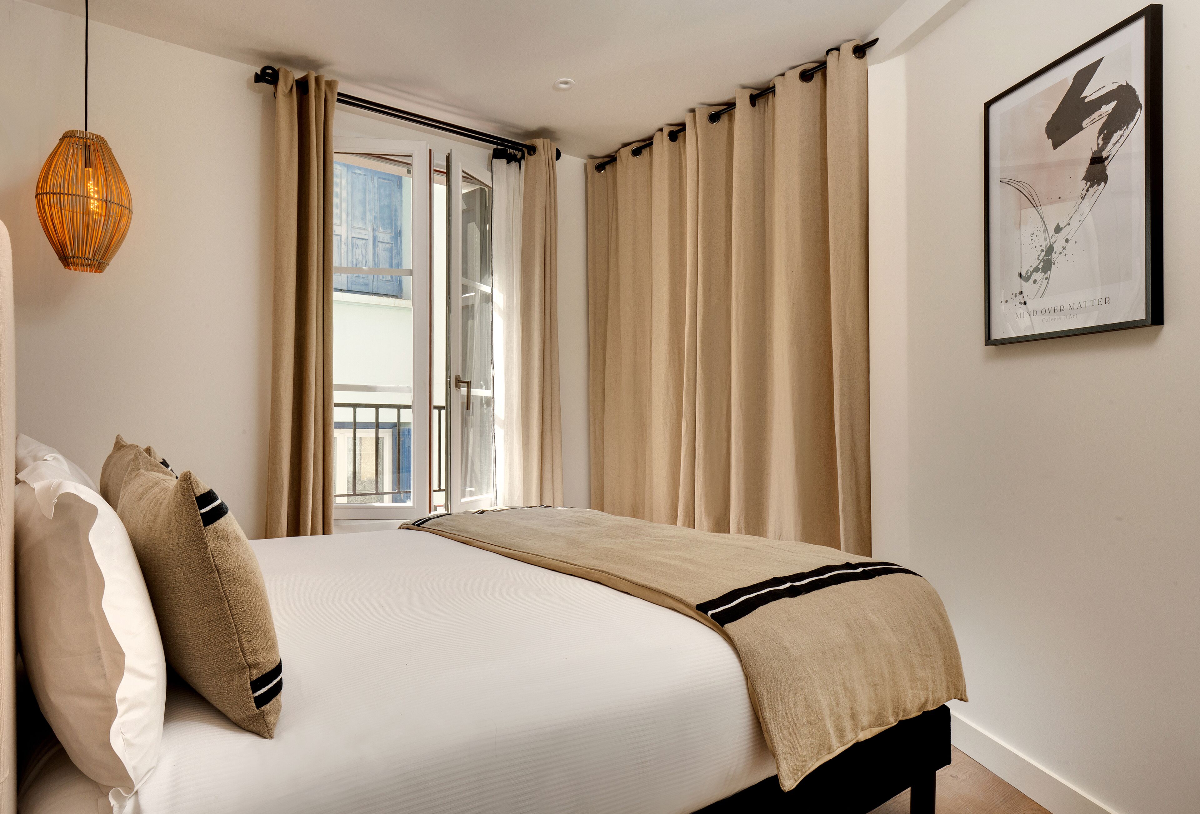 Un dormitorio con cama, cortinas color crema, una obra de arte enmarco y una lámpara colgante.