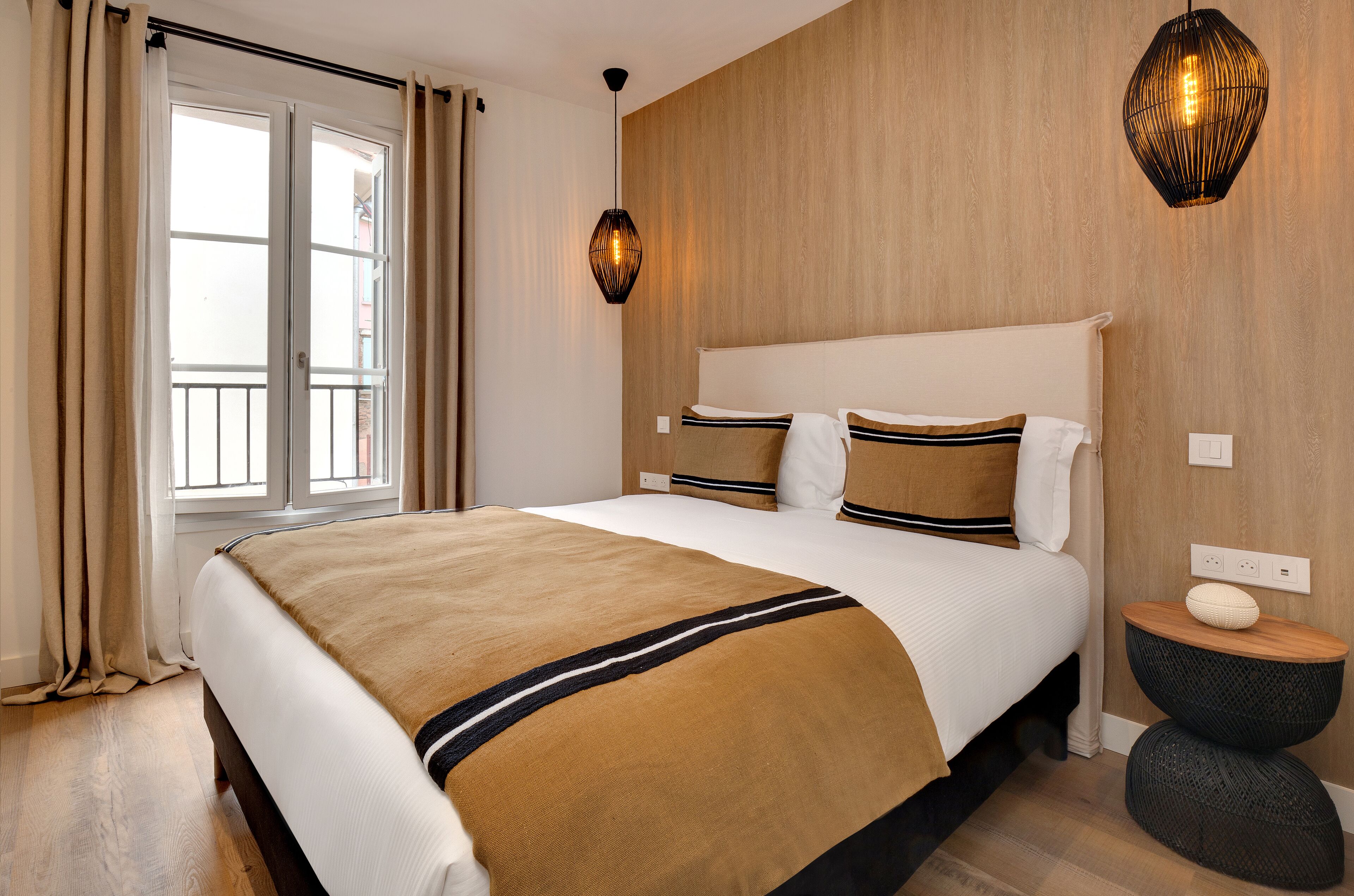 Une chambre d’hôtel avec un grand lit, une tête de lit en bois, une fenêtre avec des rideaux, un luminaire fixé au mur et une petite table de chevet en bois.