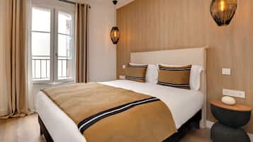 Une chambre d’hôtel avec un grand lit, une tête de lit en bois, une fenêtre avec des rideaux, un luminaire fixé au mur et une petite table de chevet en bois.
