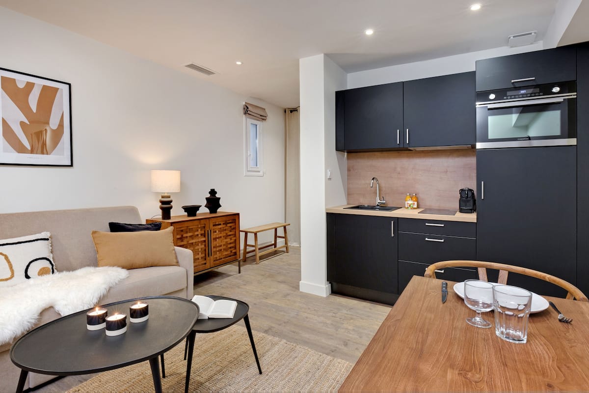 A modern kitchen with dark cabinets, a built-in oven, and a coffee maker. There is a dining table with two glasses and a knife.