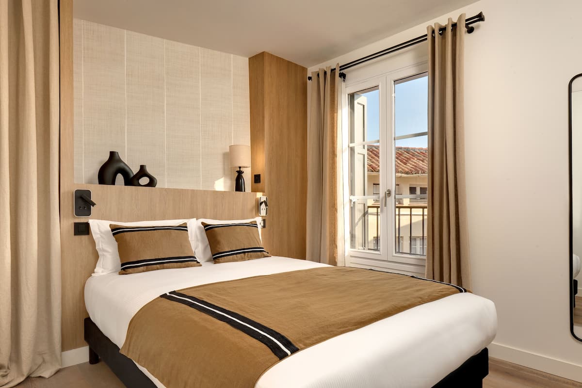A hotel room with a large bed, a wooden headboard, a window with curtains, and a view of buildings outside.
