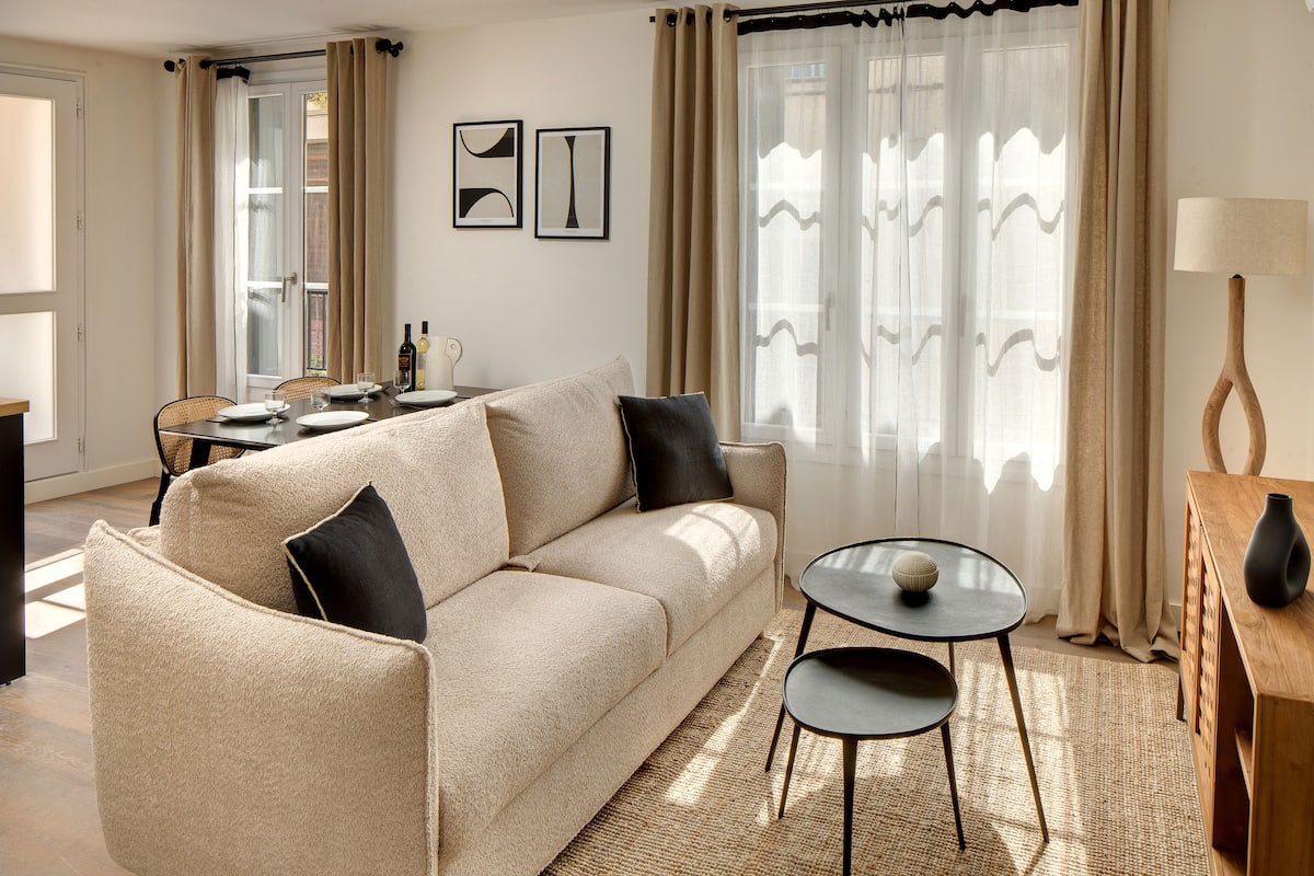 A living room with a beige sofa, a small round table, and a lamp. There is a dining table with chairs and a window with curtains.
