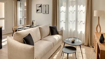Un salon avec un canapé beige, une petite table ronde et une lampe. On y trouve également une table à manger avec des chaises, ainsi qu’une fenêtre avec des rideaux.