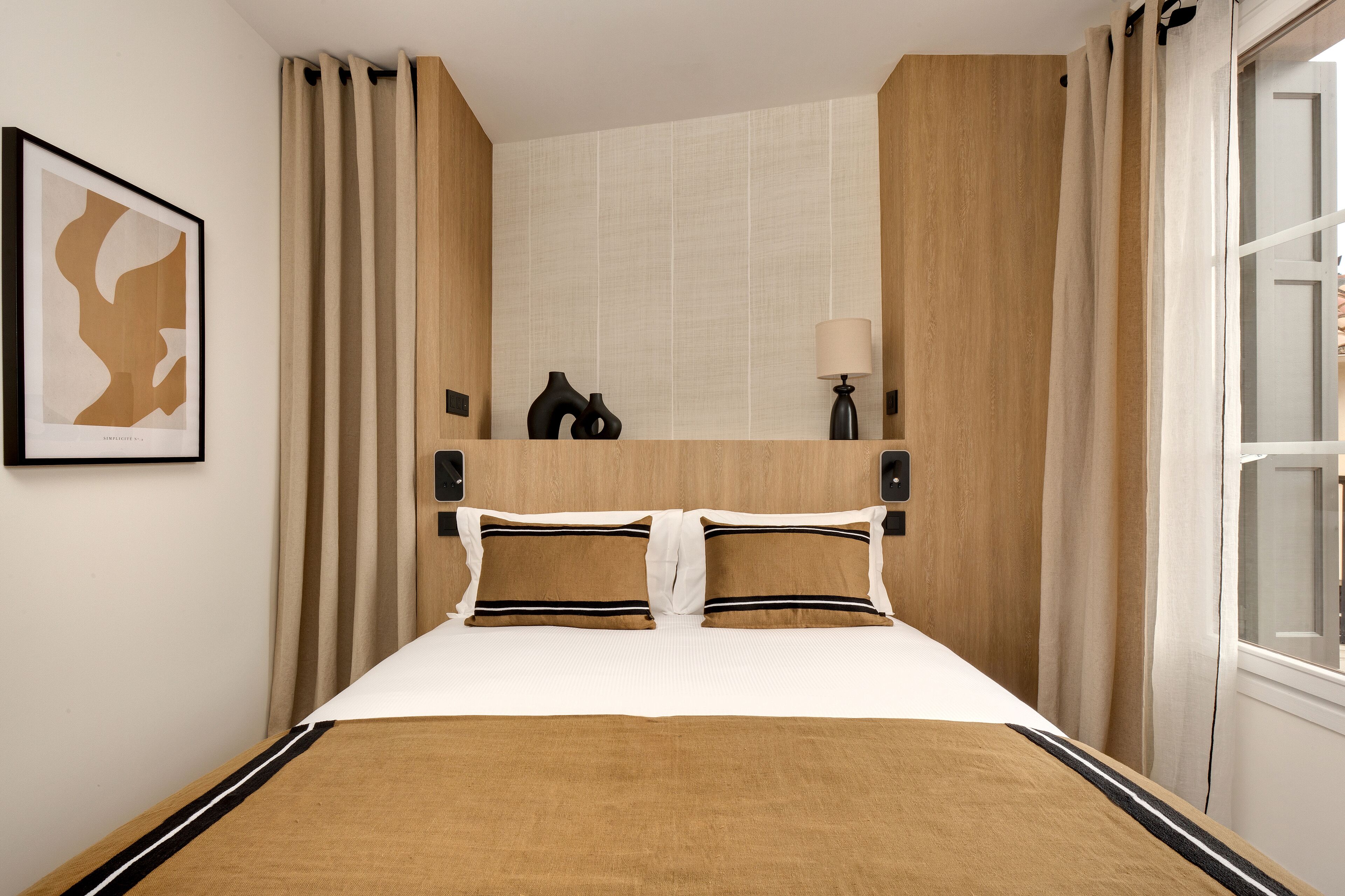 A hotel room with a large bed, beige curtains, a framed abstract artwork, a bedside lamp, and a wooden headboard.