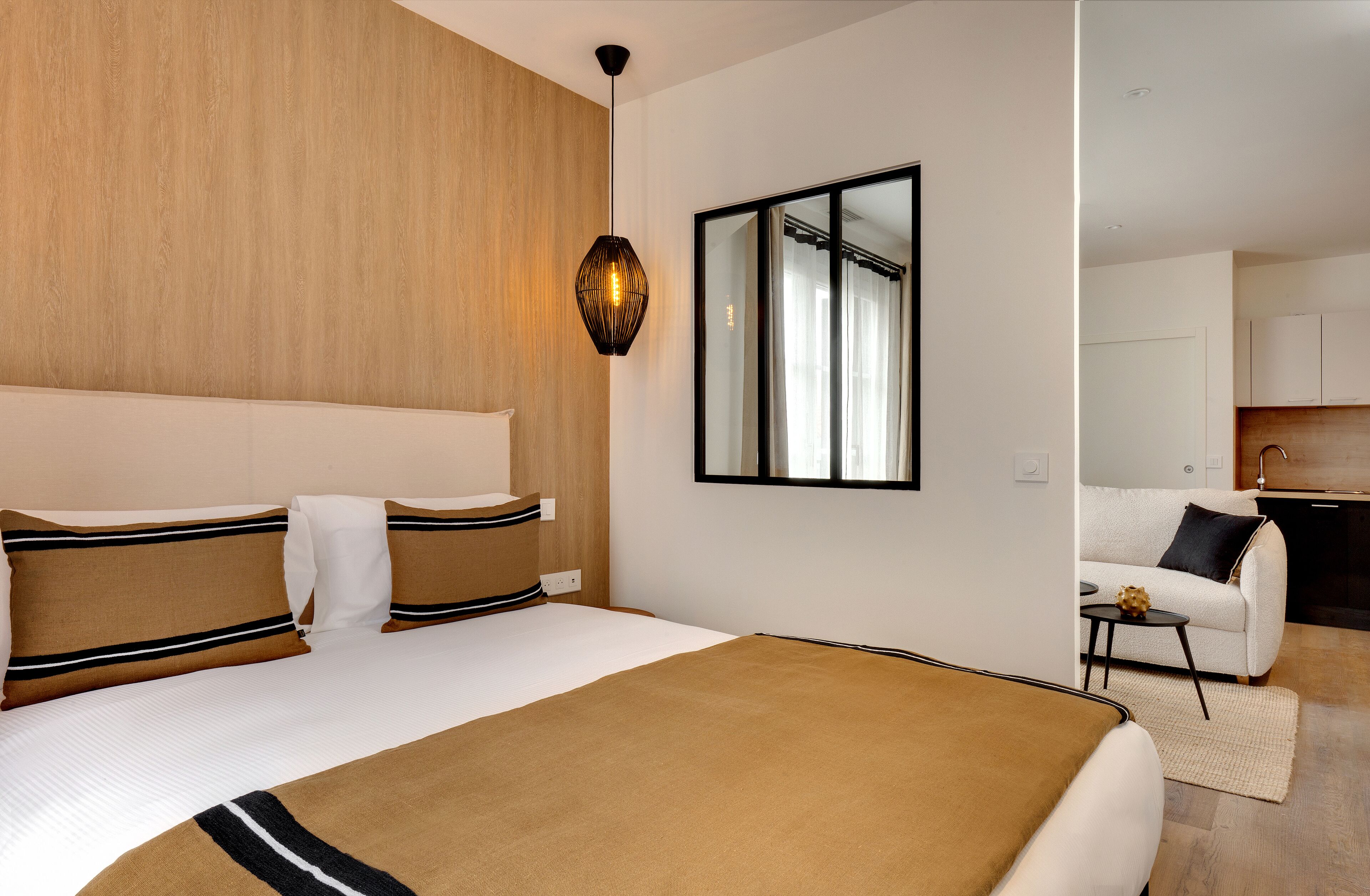 A modern hotel room with a large bed, a wooden headboard, a hanging light fixture, a mirror, and a view into the kitchen area.