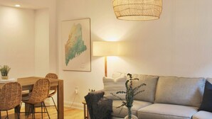 Een moderne woonkamer met een houten eettafel, rieten stoelen, een grijze bank, een rond bijzettafeltje met een vaas, en een hangende geweven lamp.