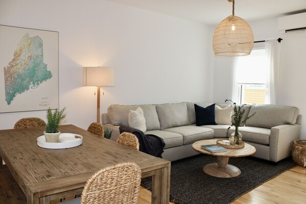 A living room with a wooden dining table, a grey sofa, wicker chairs, a round coffee table, and a hanging light fixture.