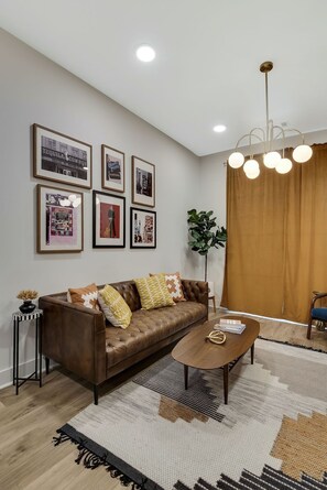 A modern living room with a brown leather sofa, a round wooden coffee table, and a large plant. - Downtown Skyline Views | Rooftop Deck | Walk to (Nashville)
