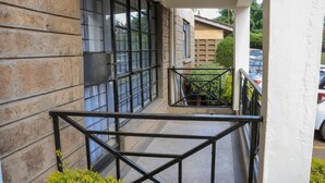 A balcony with a metal railing, a view of parked cars, and a building with large windows. - HAZINA ESTATE SOUTH B (Nairobi)