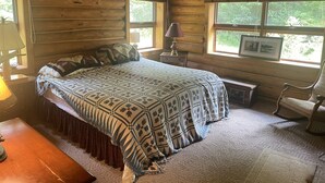 Une chambre aménagée dans un chalet en rondins, avec un grand lit, un bureau en bois, une chaise et une fenêtre donnant sur la forêt.