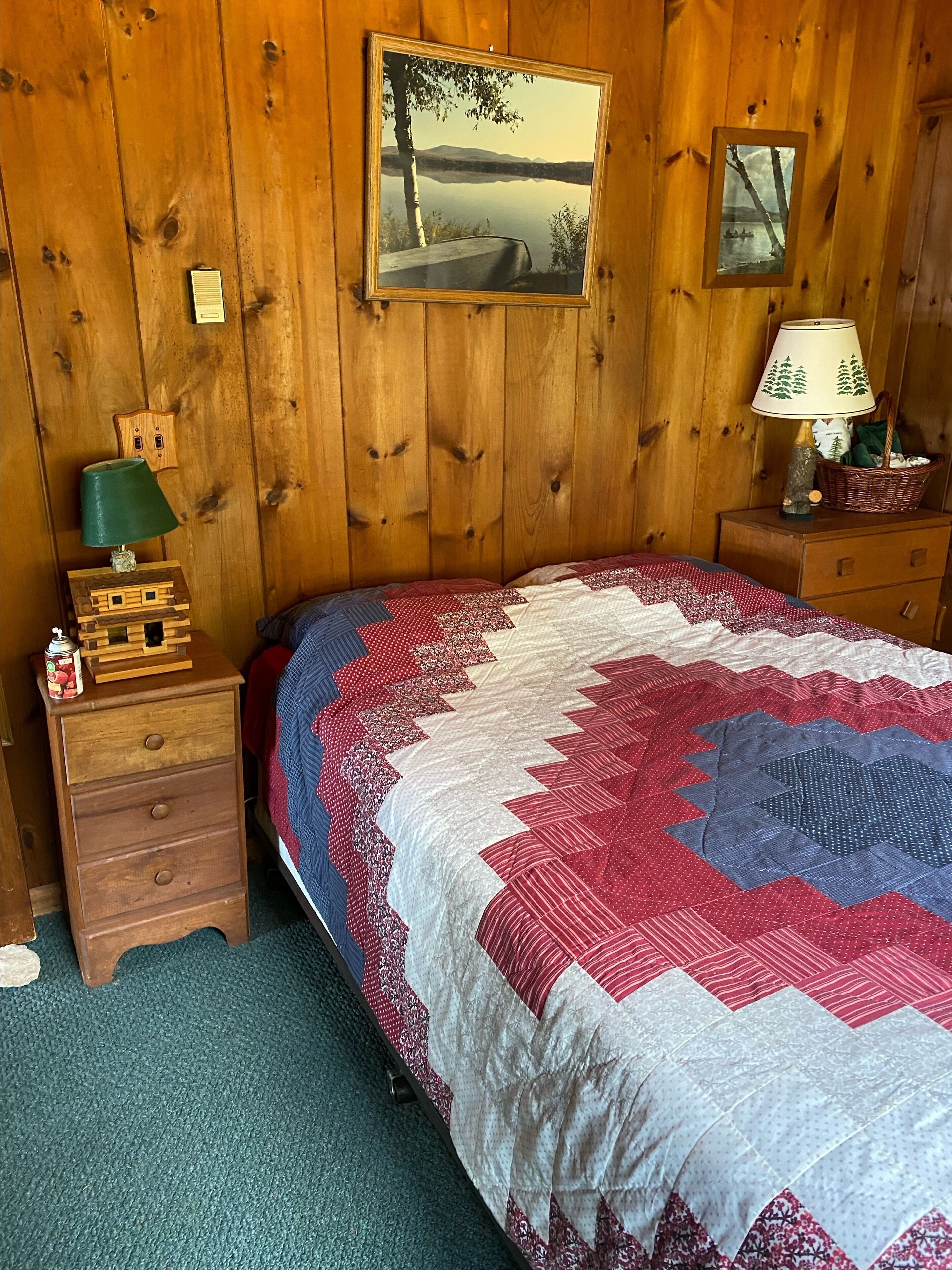 Un dormitorio de cabaña de madera con una cama grande, una mesita de noche con lámpara, un aparador y dos cuadros en la pared.