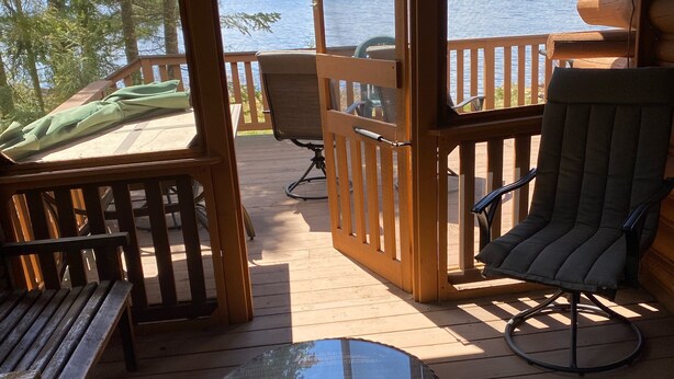 Une terrasse en bois avec une table en verre, des chaises et un banc. Une balustrade en bois entoure la terrasse, et on peut apercevoir, à travers une porte ouverte, un lac et des arbres.