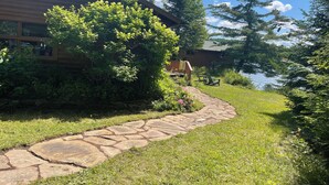 Un sentier de pierres mène à un chalet en bois situé au bord d’un lac, entouré d’une végétation luxuriante et d’arbres.