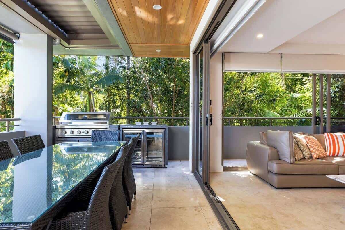 An outdoor dining area with a glass table and chairs, a grill, and a sofa with cushions.