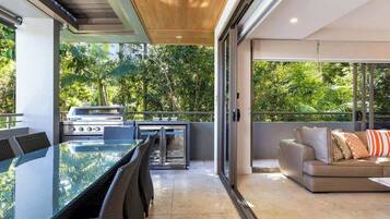 An outdoor dining area with a glass table and chairs, a grill, and a sofa with cushions.