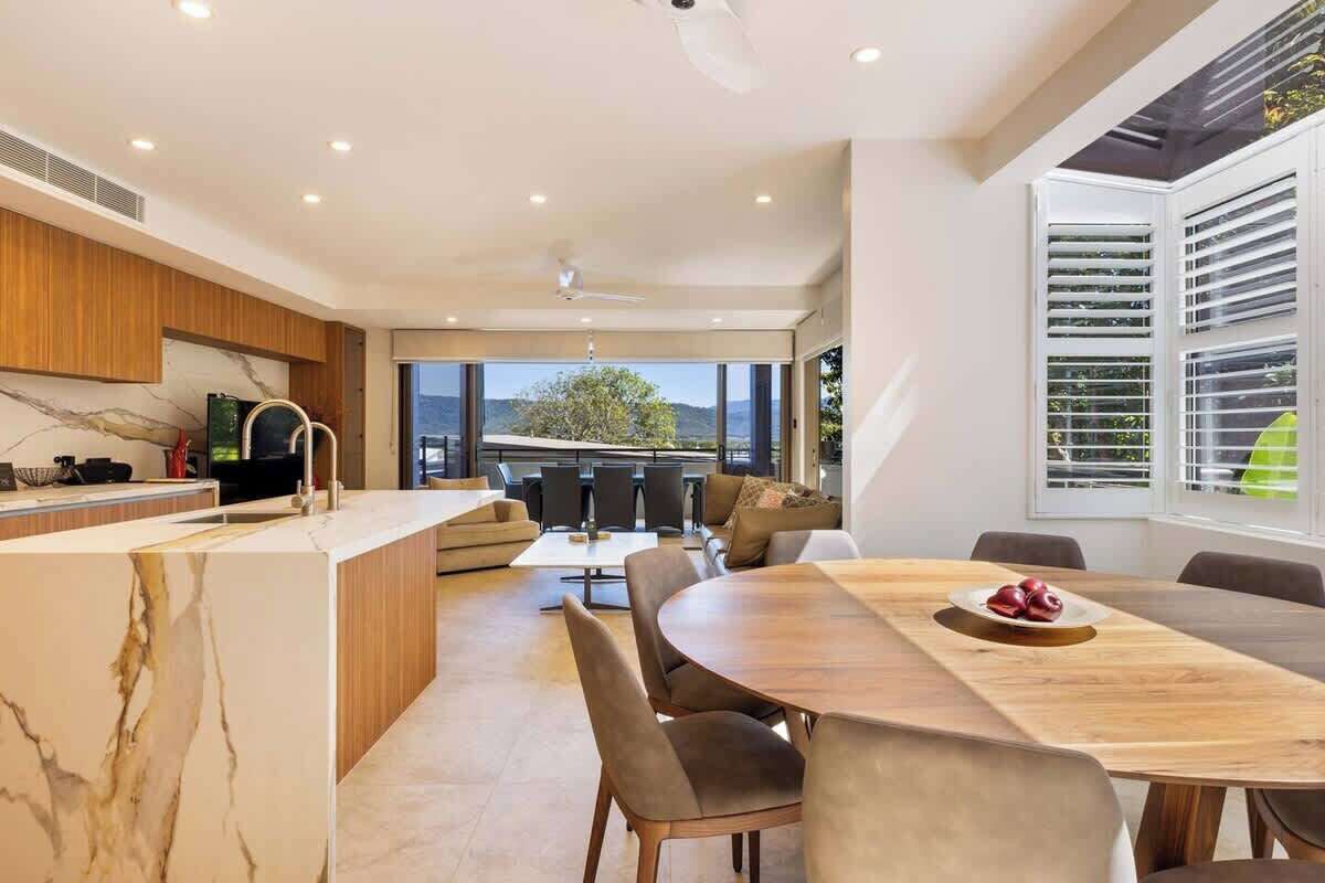 A modern kitchen with a marble countertop, a dining area with a wooden table and chairs, and large windows offering a view of the outdoors.