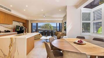 A modern kitchen with a marble countertop, a dining area with a wooden table and chairs, and large windows offering a view of the outdoors.